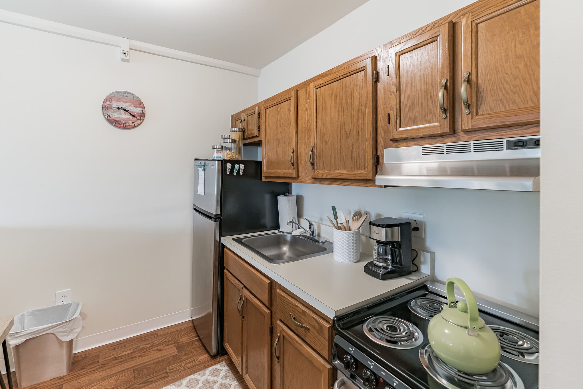 - Grace Management A kitchen with an American flag-patterned circular wall clock, light-brown cabinets, a fridge, sink, a coffee maker, and a green kettle on the gas stove at Shorehaven, a top choice for senior living near Warren.