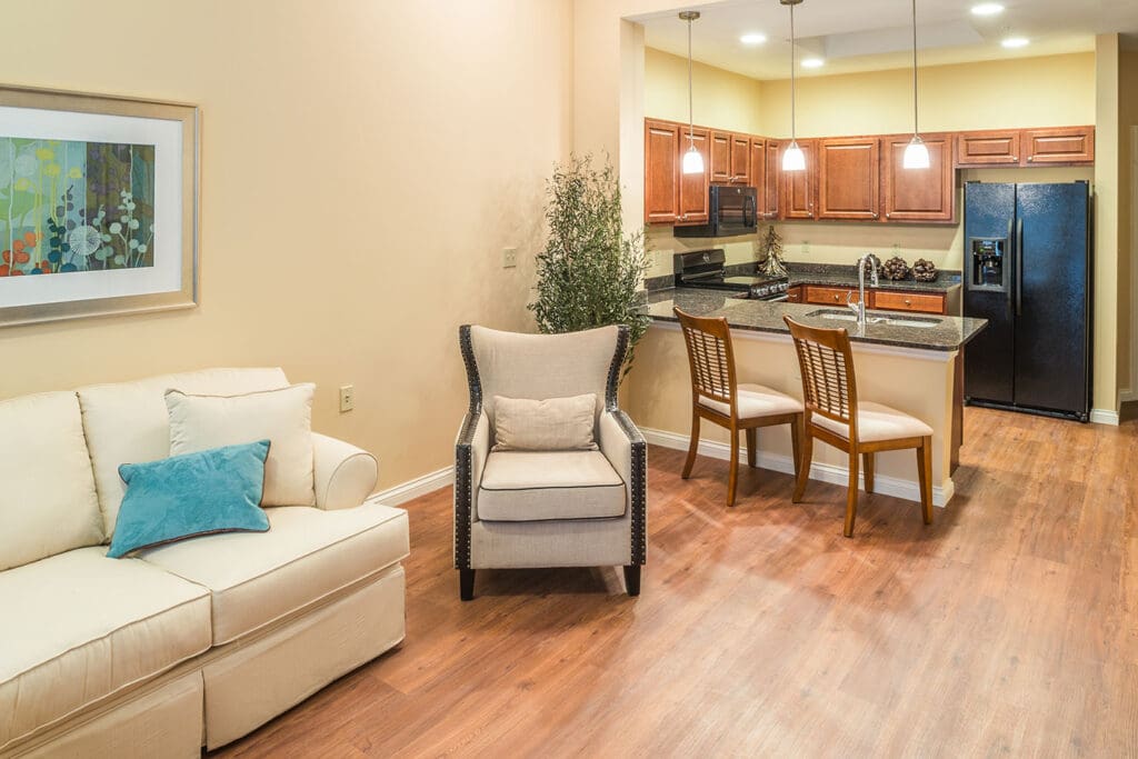 - Grace Management A living room area with a beige couch, beige sofa chairs, with access to the kitchen with light brown cupboards, gray marbled counters, bar stools, a sink, and a black fridge.