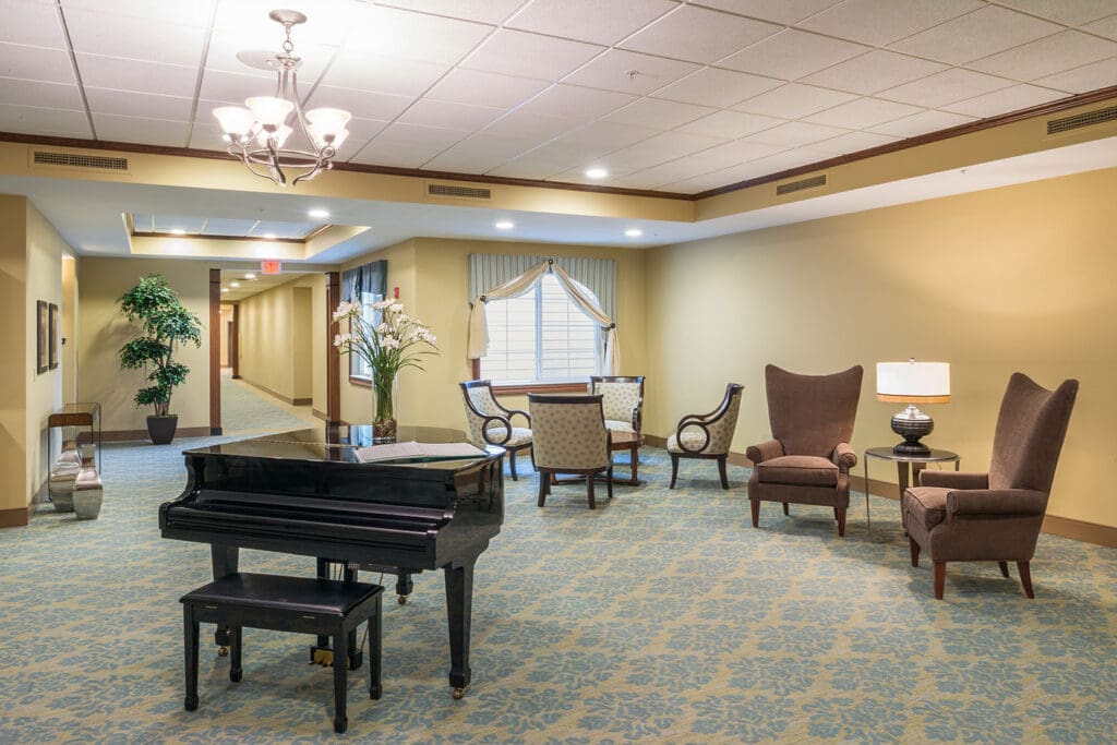 - Grace Management Second Floor Neighborhood sitting area with a piano table, a vase with white flowers in it, sofa chairs around small coffee tables.