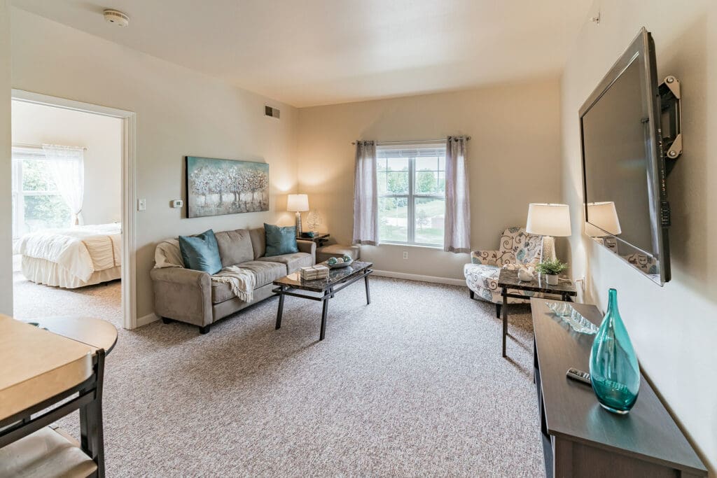 - Grace Management A living room with a gray couch and blue throw pillows on it, a coffee table in front of it, a sofa chair, and a wall-mounted TV above a TV stand at Town Village Sterling Heights, a senior living community near Detroit.