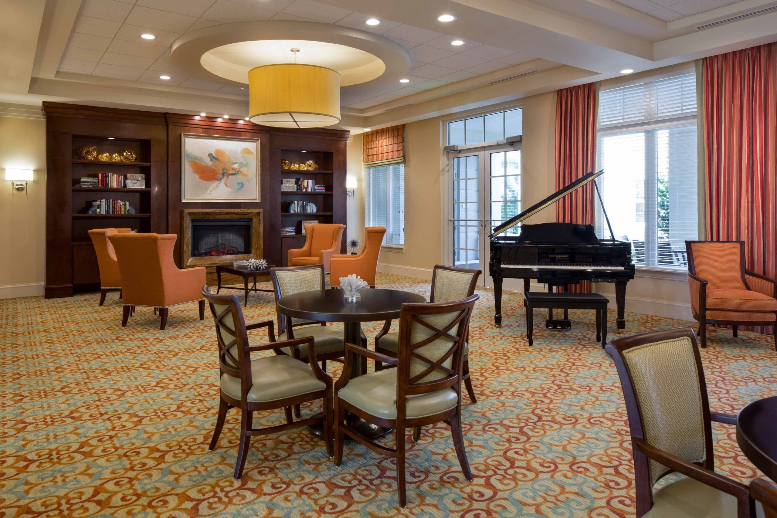 A carpeted lobby area with orange curtains, a piano, tables, and chairs for sitting and lounging at The Summit of Lakewood Ranch.