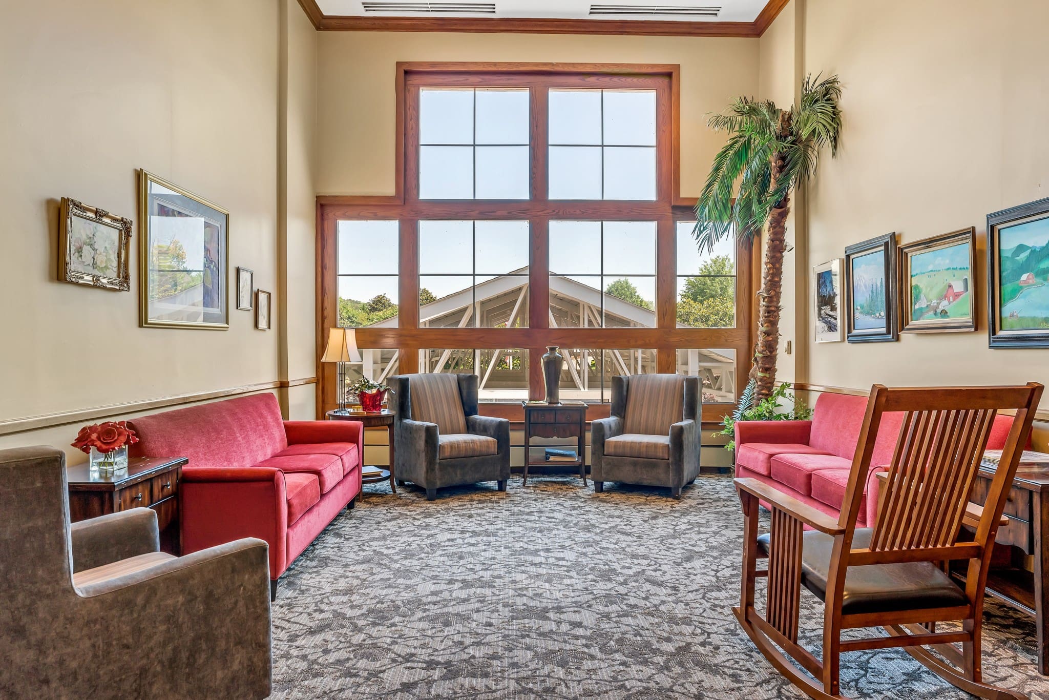 A sitting area with two red couches facing one another, two sofa chairs against a ceiling-high window with a side table between them and a lamp on it, a brown sofa chair, and a rocking chair at Town Village Vestavia Hills, a senior living community near Homewood, AL.