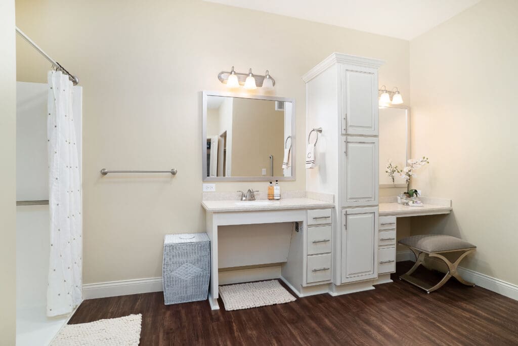 - Grace Management A photo of the bathroom with a sink, two mirrors, a vanity table and chair, and a walk-in shower at Celebration Village Forsyth, a senior living community near Cumming, GA