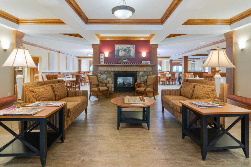 A furnished community lounge with fireplace and seating near the dining area at Dirigo Pines, a senior living community near Brewer, Maine.