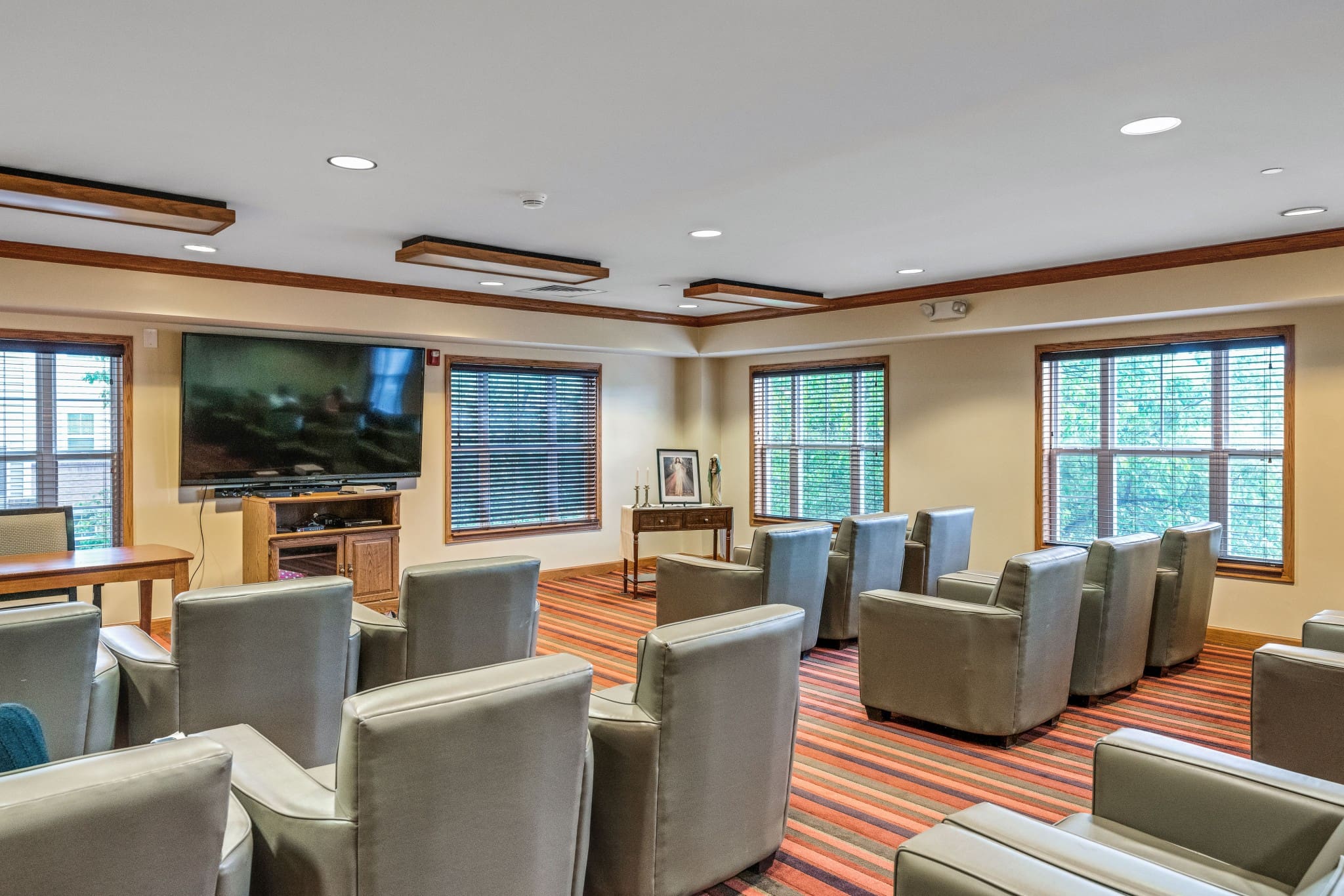 - Grace Management A movie theater room with rows of gray sofa chairs facing a TV at Town Village Sterling Heights, a senior living community near Clinton Township, MI.
