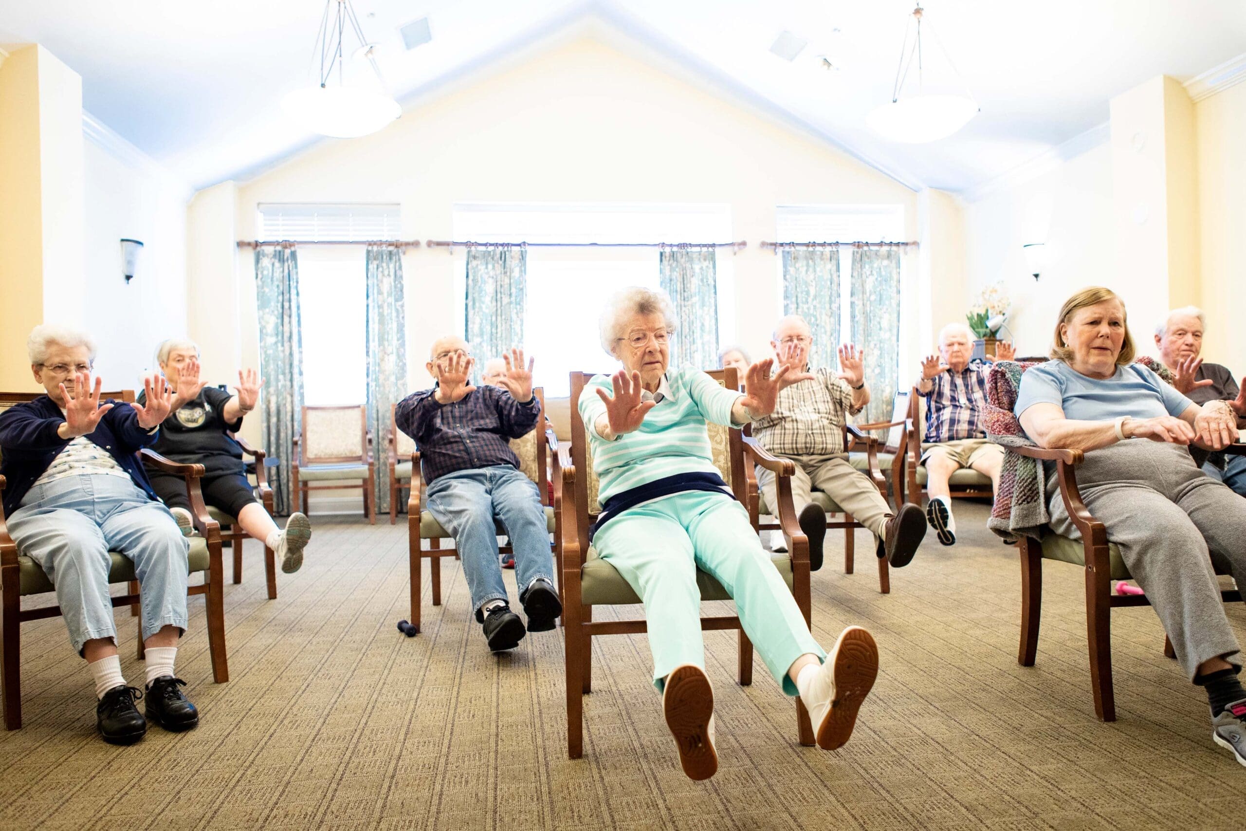 - Grace Management A group of white seniors are sitting on chairs that are spread our across the room, stretching their arms and feet forward.