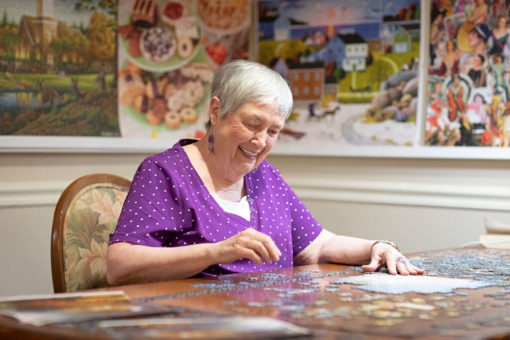 An older white woman wearing a purple dress with small white polka dots is smiling as she's sitting at the table and putting together a puzzle at Portside at Grande Dunes, a senior living community near Murrells Inlet, SC.