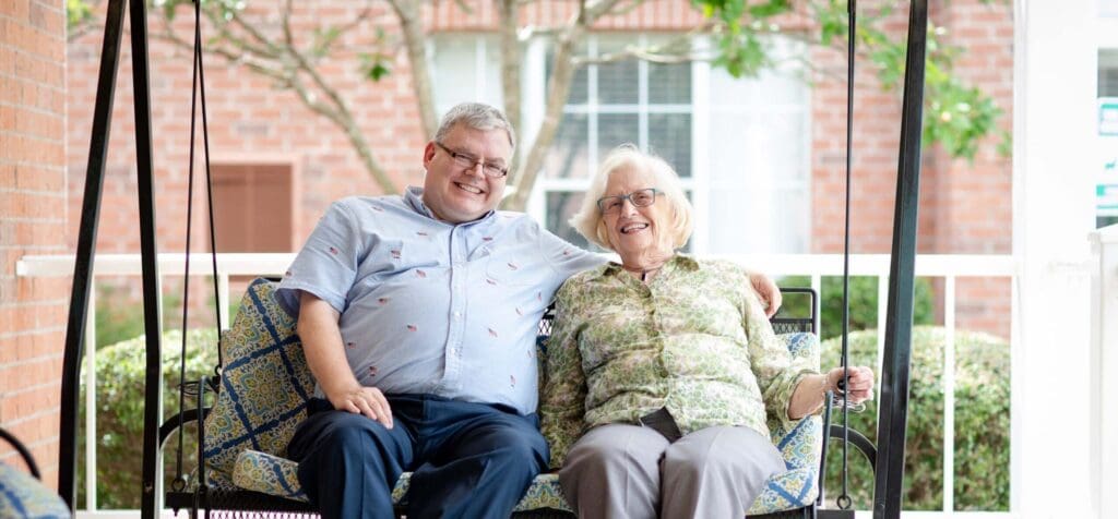A middle-aged white man is sitting on the swing next to an older white woman with his hand on her shoulder at Portside at Grande Dunes, a senior living community near Conway, SC.