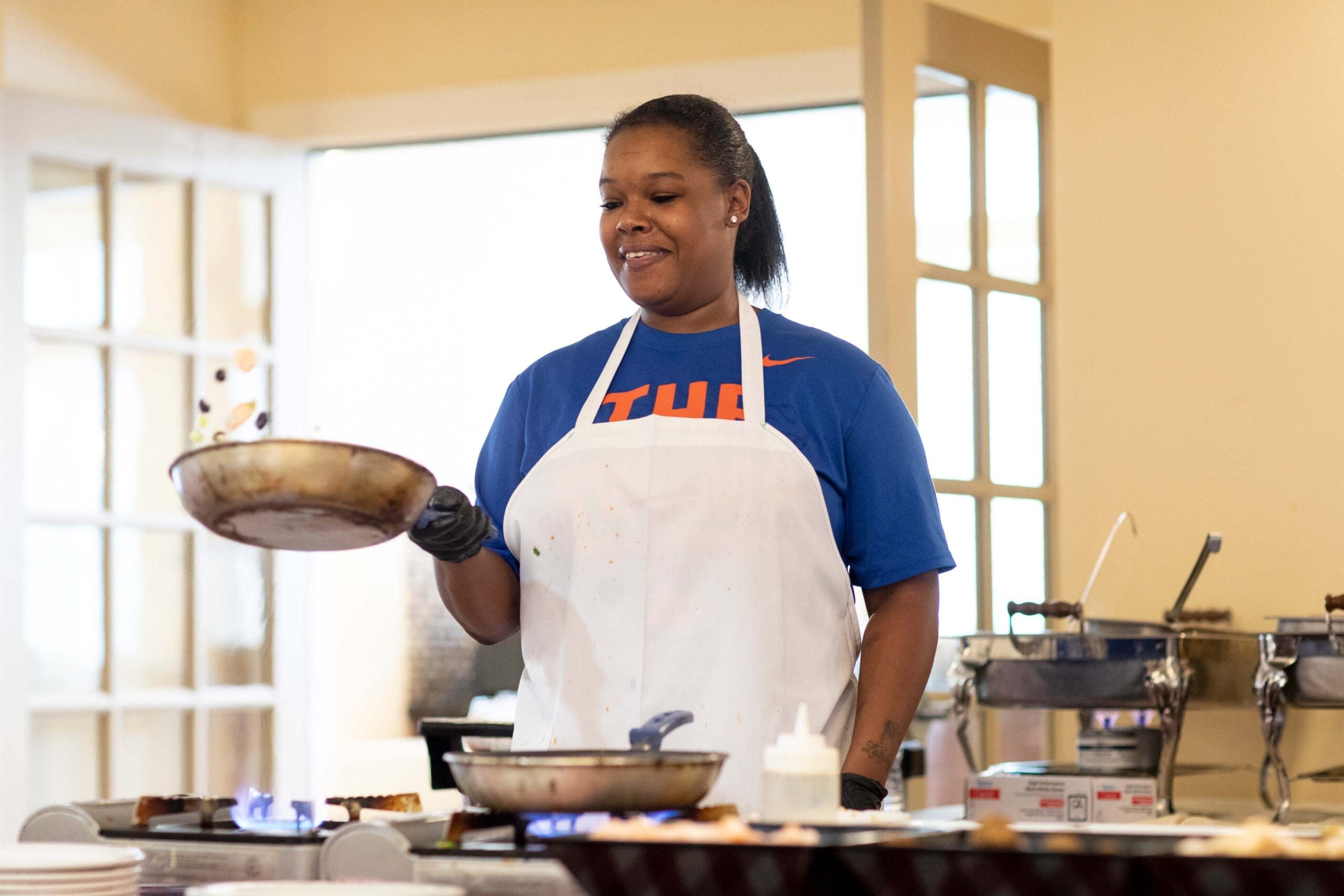 A middle-aged black woman wearing a white apron is standing in front of the stove with a frying pan in her hand, tossing vegetables up in the air.