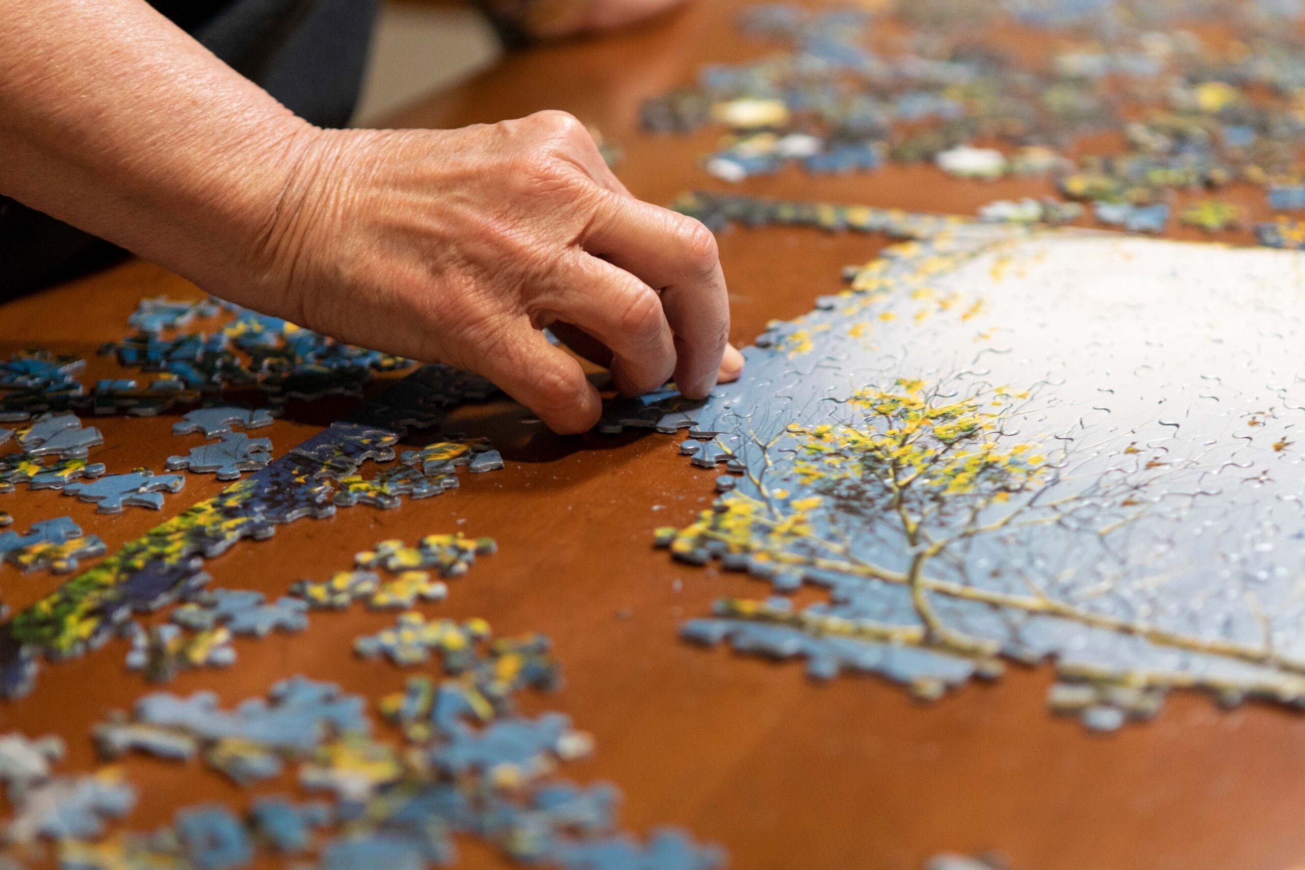 A senior person's hand is putting a puzzle piece into place on a wooden table at Parkrose Estates, a premier senior living community near Bridgeport, NY.