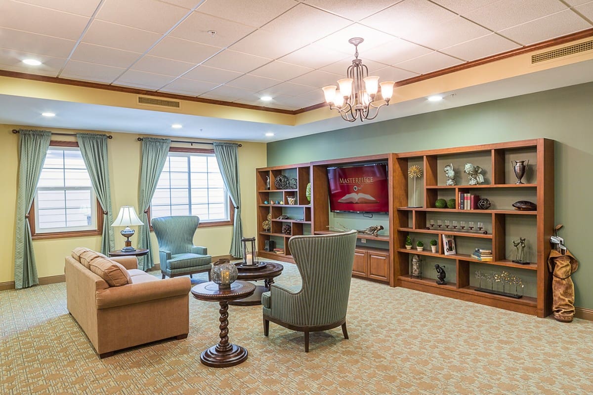 - Grace Management Grand neighborhood first floor sitting area with a light brown couch, light blue sofa chairs, side tables with lamps on them, and a wall-length shelf and a TV.