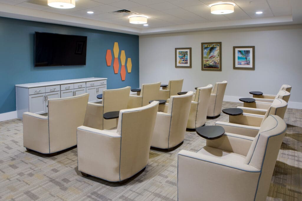 - Grace Management A movie theater room with three rows of beige sofa chairs facing a TV screen at Heritage Oaks of Englewood, a senior living community near Venice, FL.