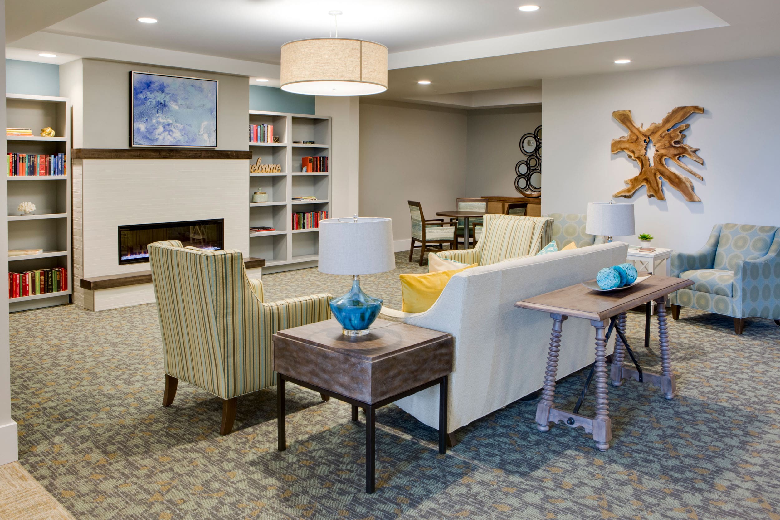 - Grace Management A living room are with a white couch and throw pillows on it, sofa chairs next to it with side tables and lamps on it, located across a digital fireplace that's underneath a wall painting, which is next to two bookshelves with books on them at Heritage Oaks of Englewood, a senior living community near Venice, FL.