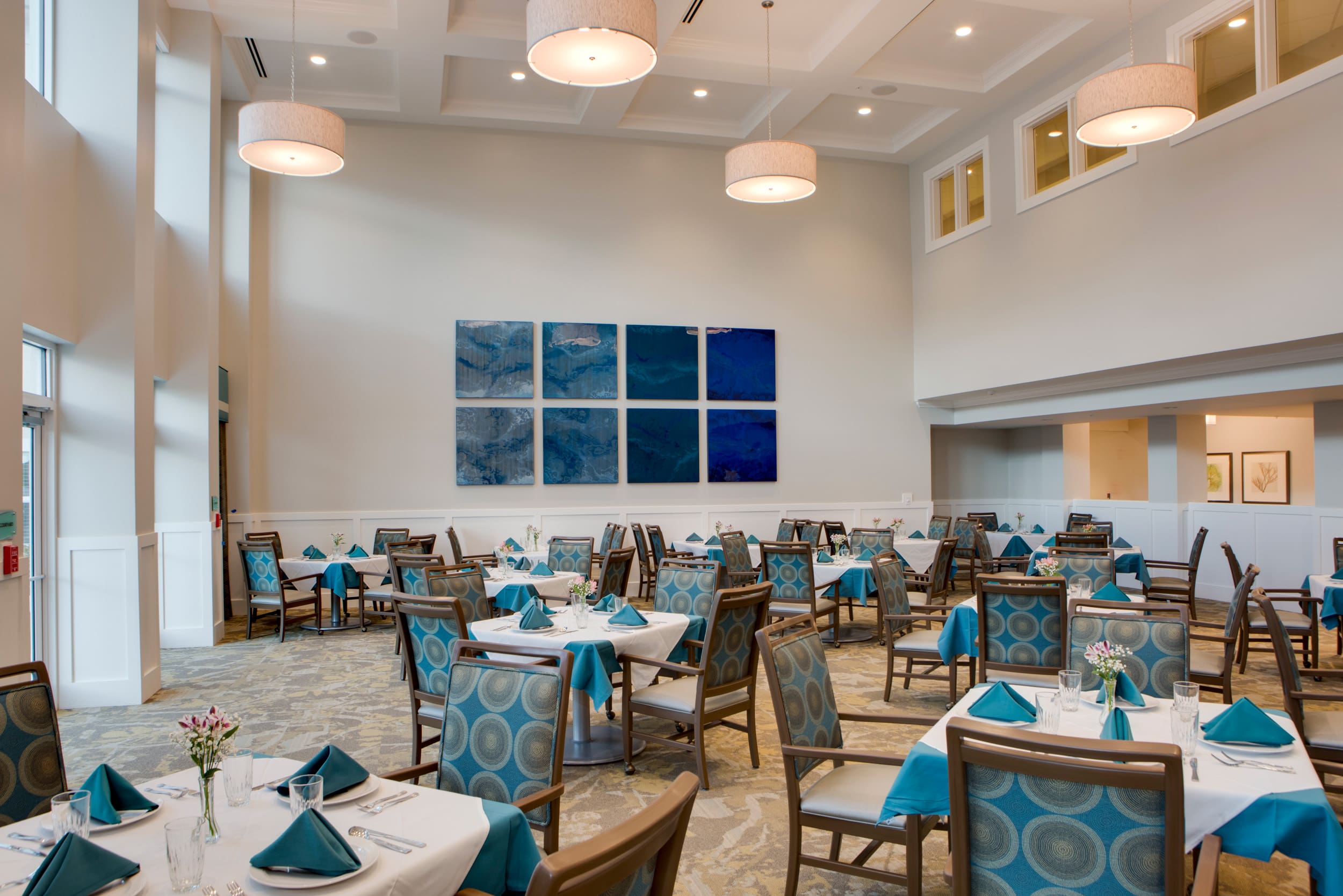 A dining area with square tables covered in blue and white table cloths with plates, glasses, silverware, and blue napkins on them, surrounded by blue and brown patterned chairs at Heritage Oaks of Englewood, a senior living community near North Port, FL.
