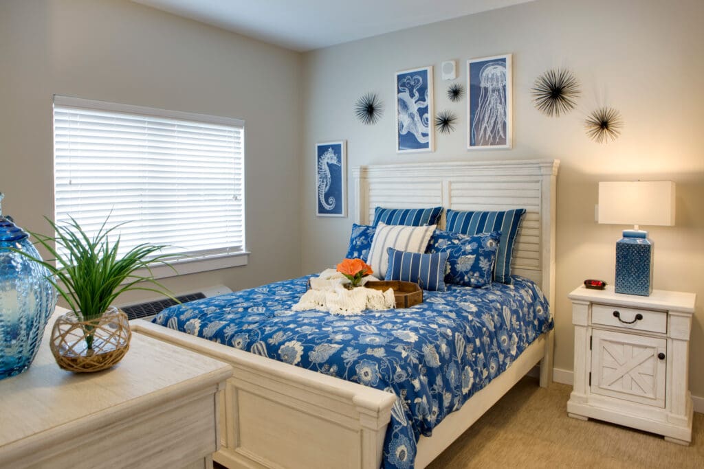 A bedroom with a Queen-sized bed with blue bedding and throw pillows underneath three wall paintings of sea creatures, with a white bedside table to the left of the bed and a lamp on top of it at Heritage Oaks of Englewood, a senior living community near North Port, FL.