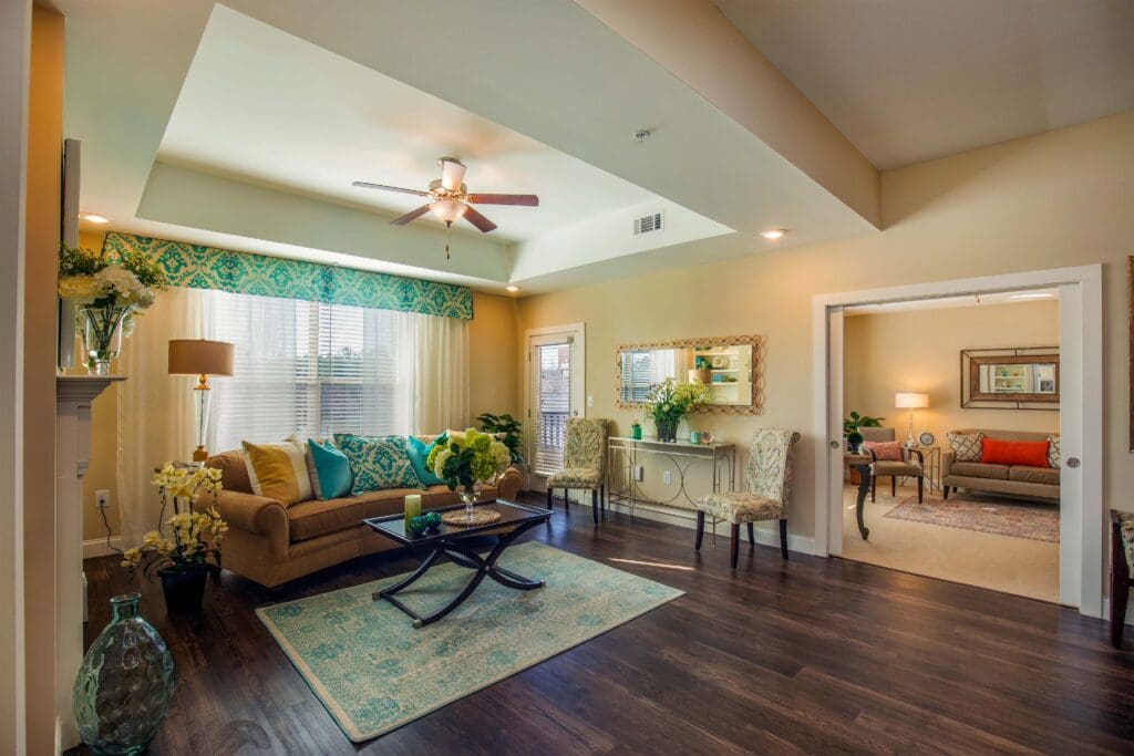 - Grace Management A living room with a mustard couch, orange and blue throw pillows, a coffee table sitting on top of a blue and white carpet, a bouquet of yellow flowers on top of the table and to the left of the couch, and two white and blue patterned chairs to the right of the couch. The sliding doors on the wall reveal another living room.