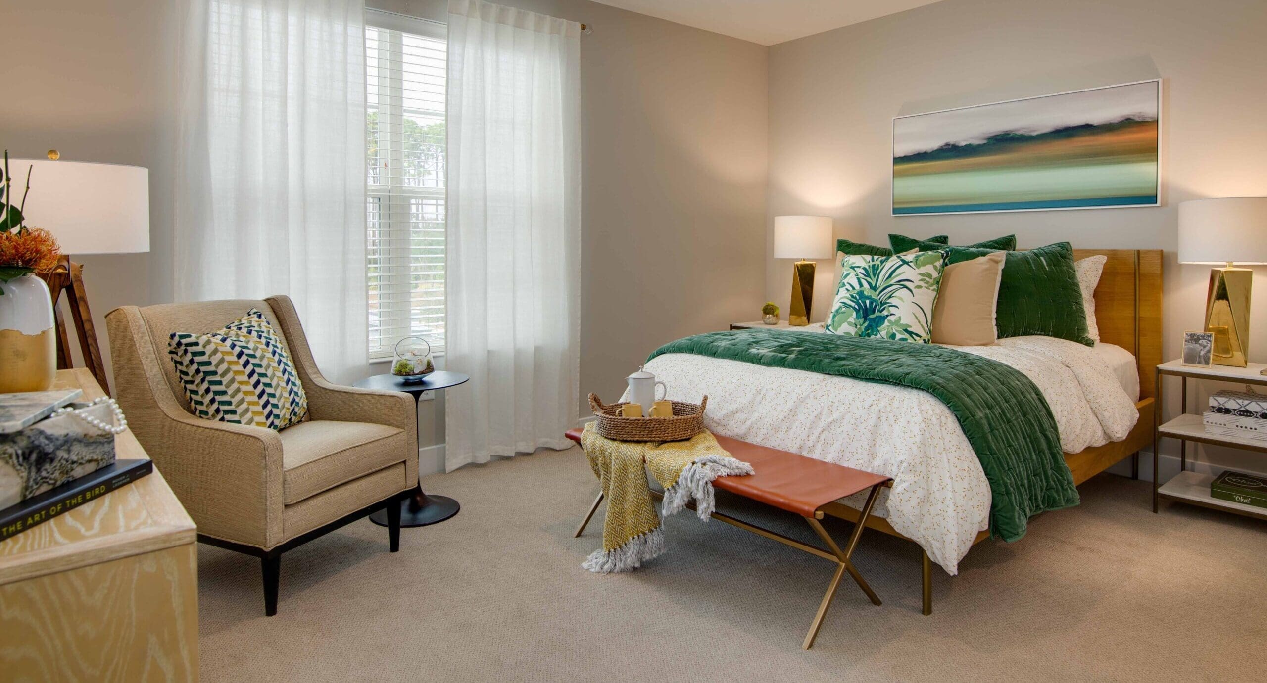 A bedroom with a made-up bed with white sheets, green throw blanket, green throw pillows, a landscape painting above the bed, two bedside tables with lamps on each side, a sitting sofa chair across the room, and an orange footstool with a yellow throw blanket on it. Portside at Grande Dunes, a senior living community near Murrells Inlet, SC.