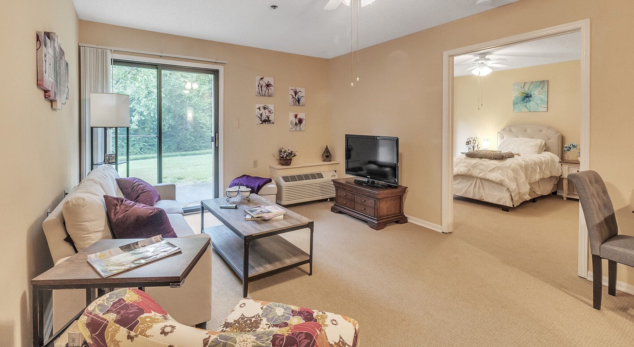 - Grace Management A living room with a gray couch and purple throw pillows, a coffee table, a TV, and a bedroom seen through the opening in the wall with a white bed at Northgate Park.