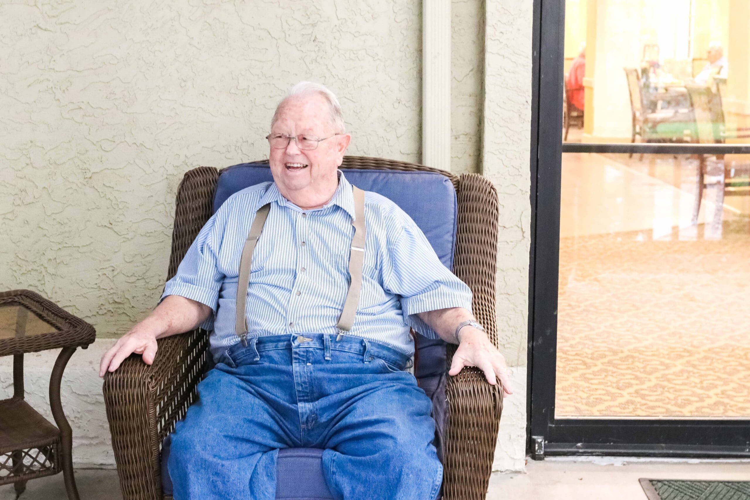 - Grace Management Smiling resident sitting in wicker chair on the patio at Sedgwick Plaza, a top choice for senior living.