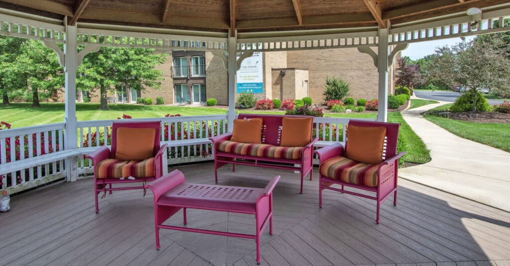 - Grace Management A gazebo with a pink couch and two pink sofa chairs with orange throw pillows on them and a pink coffee table in front of them at Atrium of Belleville.