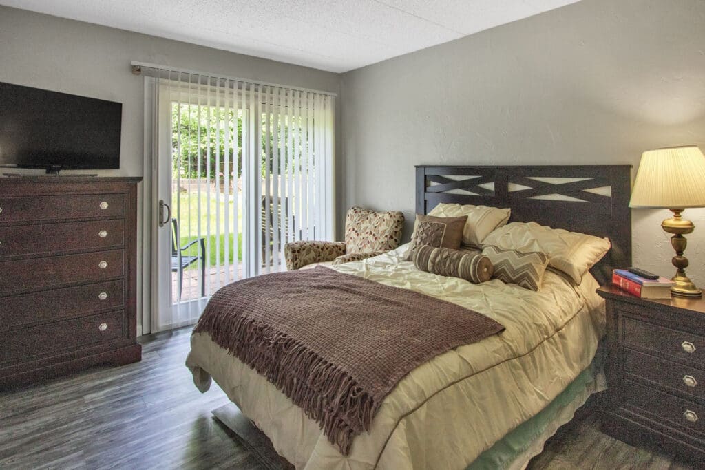 - Grace Management A bedroom with a made-up bed with gold sheets and pillows and a brown throw blanket, a bedside table with a lamp and books on it, a sofa chair, a dresser, and a TV on top of it at the atrium of Belleville.