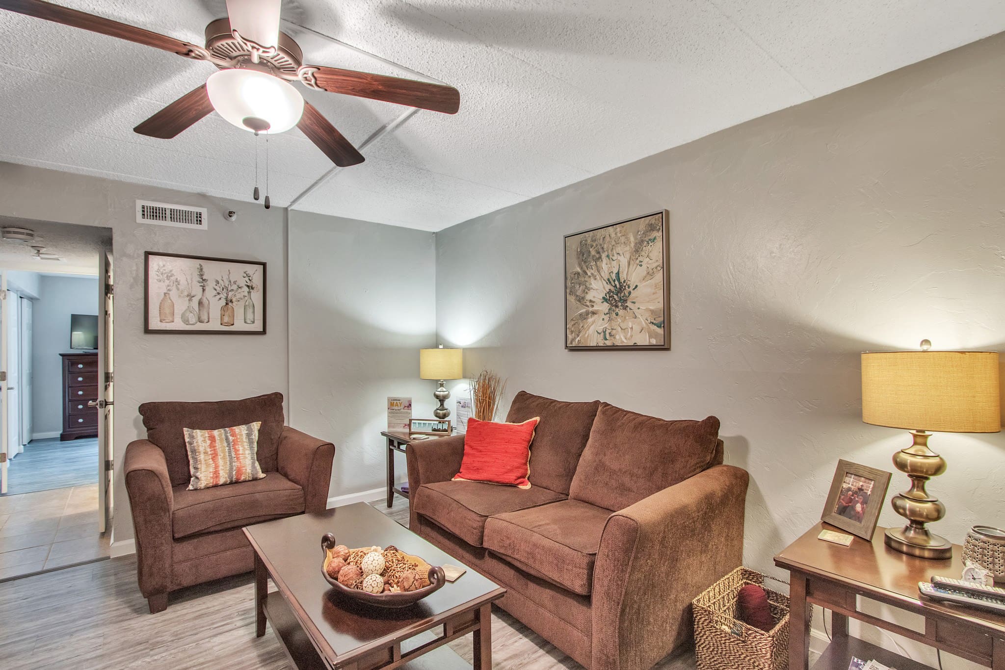 - Grace Management A living room with a brown couch and sofa chair, a coffee table, a ceiling fan, and a side table with a lamp and a framed photograph at The Atrium of Belleville.