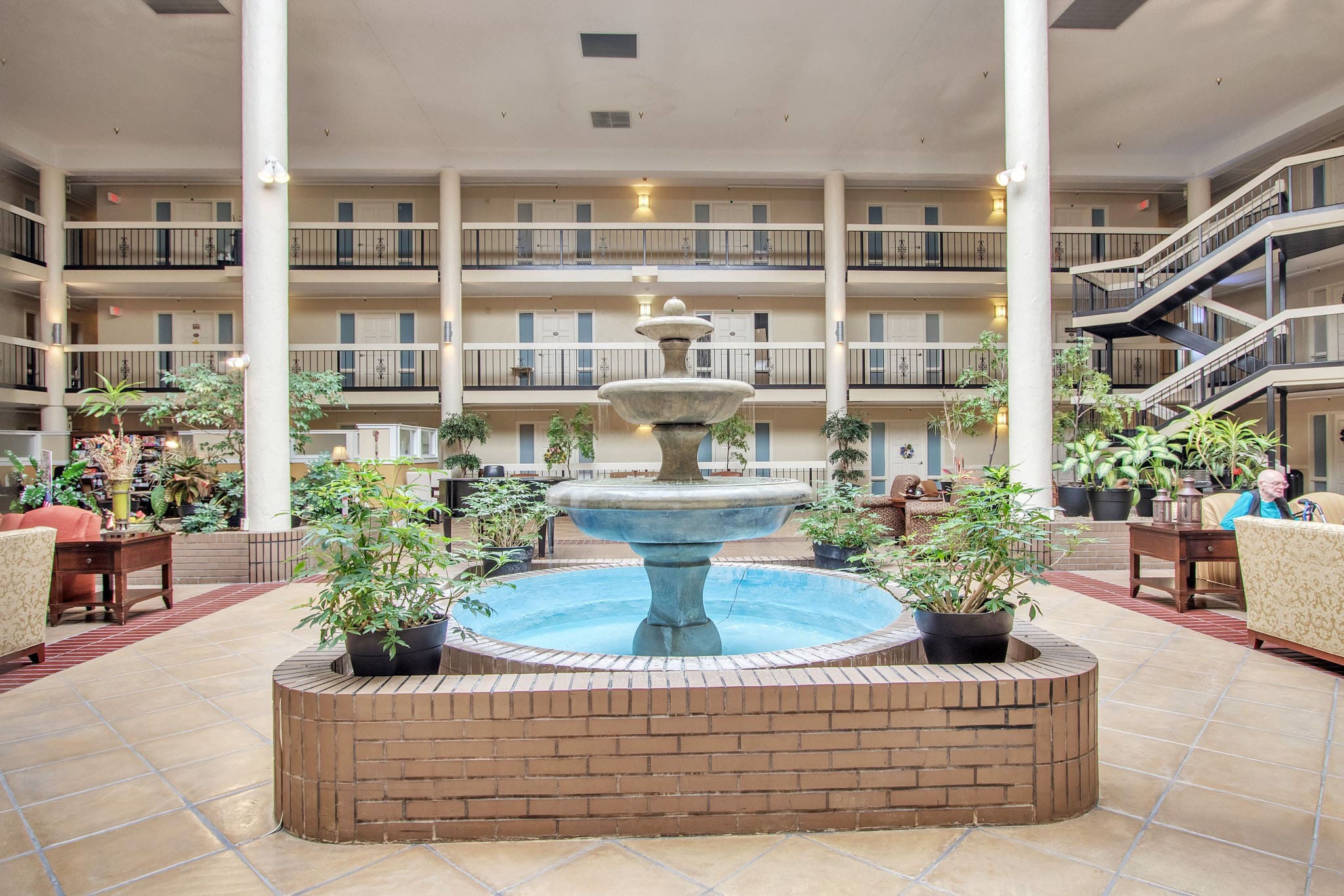 - Grace Management Fountain in the middle of The Atrium of Belleville lobby, with potted plants surrounding it.