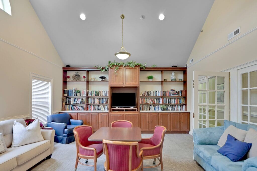- Grace Management An indoor sitting area with white and blue couches, a blue sofa chair, a brown table with pink cushioned chairs around it, all located in front of a long bookshelf full of books and a TV.