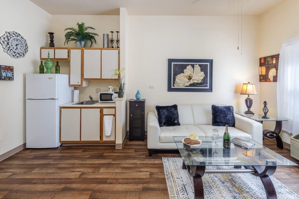 - Grace Management Inside of apartment showing kitchen area with refrigerator and microwave, living area with cozy couch, a coffee table with wine, book, and a snack. Artwork hangs on the walls. At the Vista Pointe at Mira Loma.