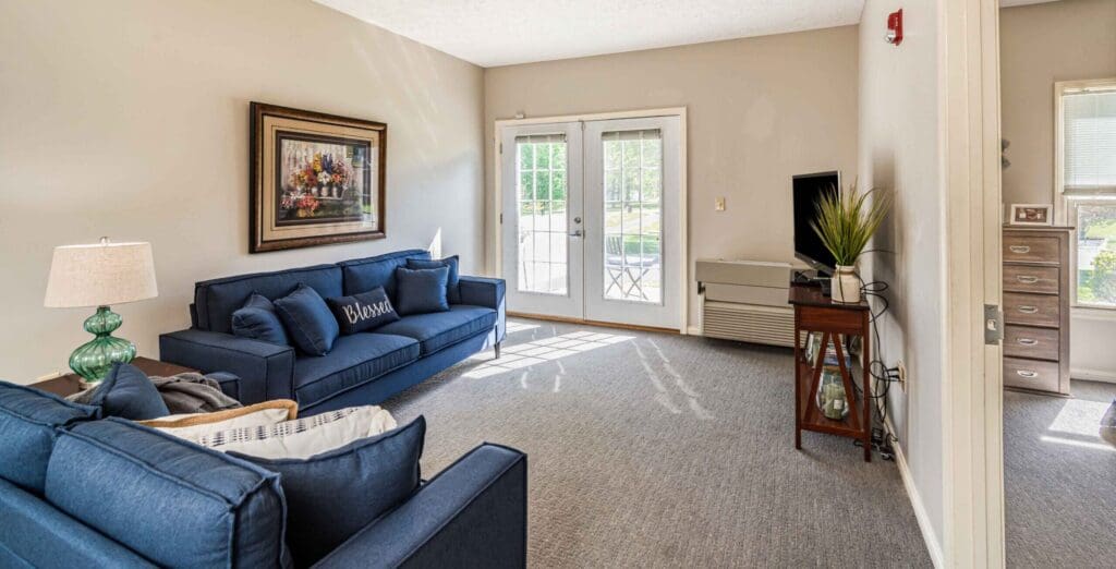 - Grace Management Interior image of a resident's living room with comfortable furniture and colorful artwork. There is a double door leading out to a patio space on the far wall.