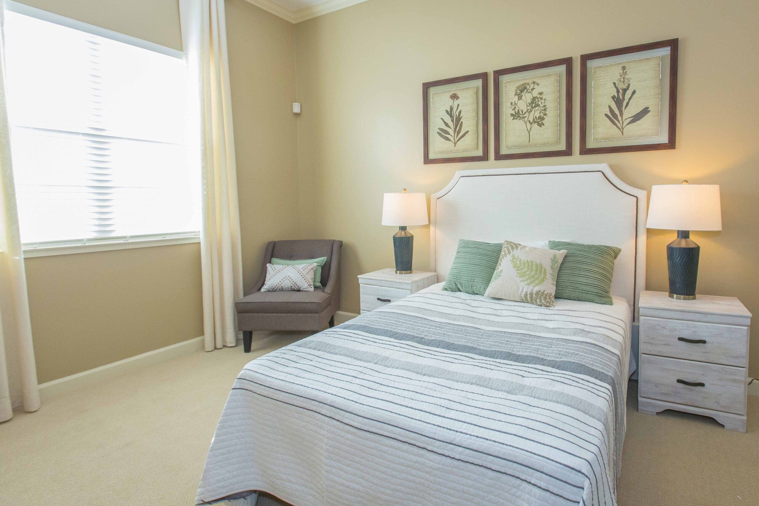 A bedroom with a made-up bed with white sheets, green pillows, two bedside tables with lamps on them, and a gray sofa chair with throw pillows in the corner.