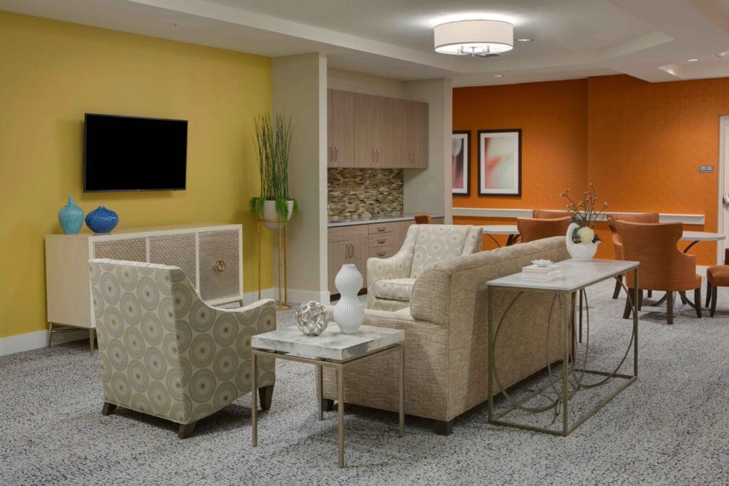 A sitting area with yellow and orange walls, a TV mounted to the wall, a beige couch, beige sofa chairs, and side tables with vases on them at The Summit of Winter Garden, a senior living community in Orange County.