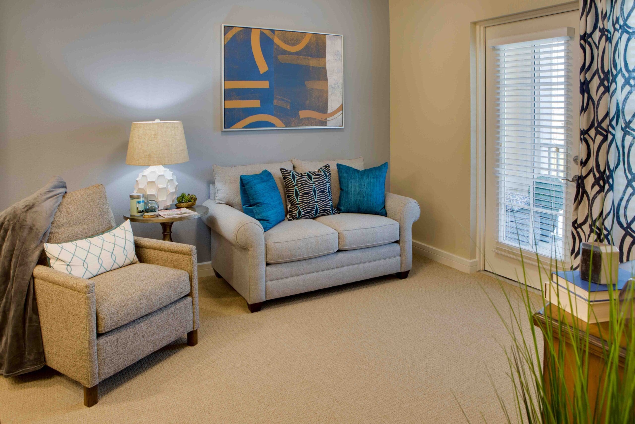 A living room with a gray couch with blue throw pillows, a beige sofa chair, a side table with a lamp on it, and a window leading to the balcony with blue curtains at The Summit of Winter Garden, an independent and assisted living community near Orlando.