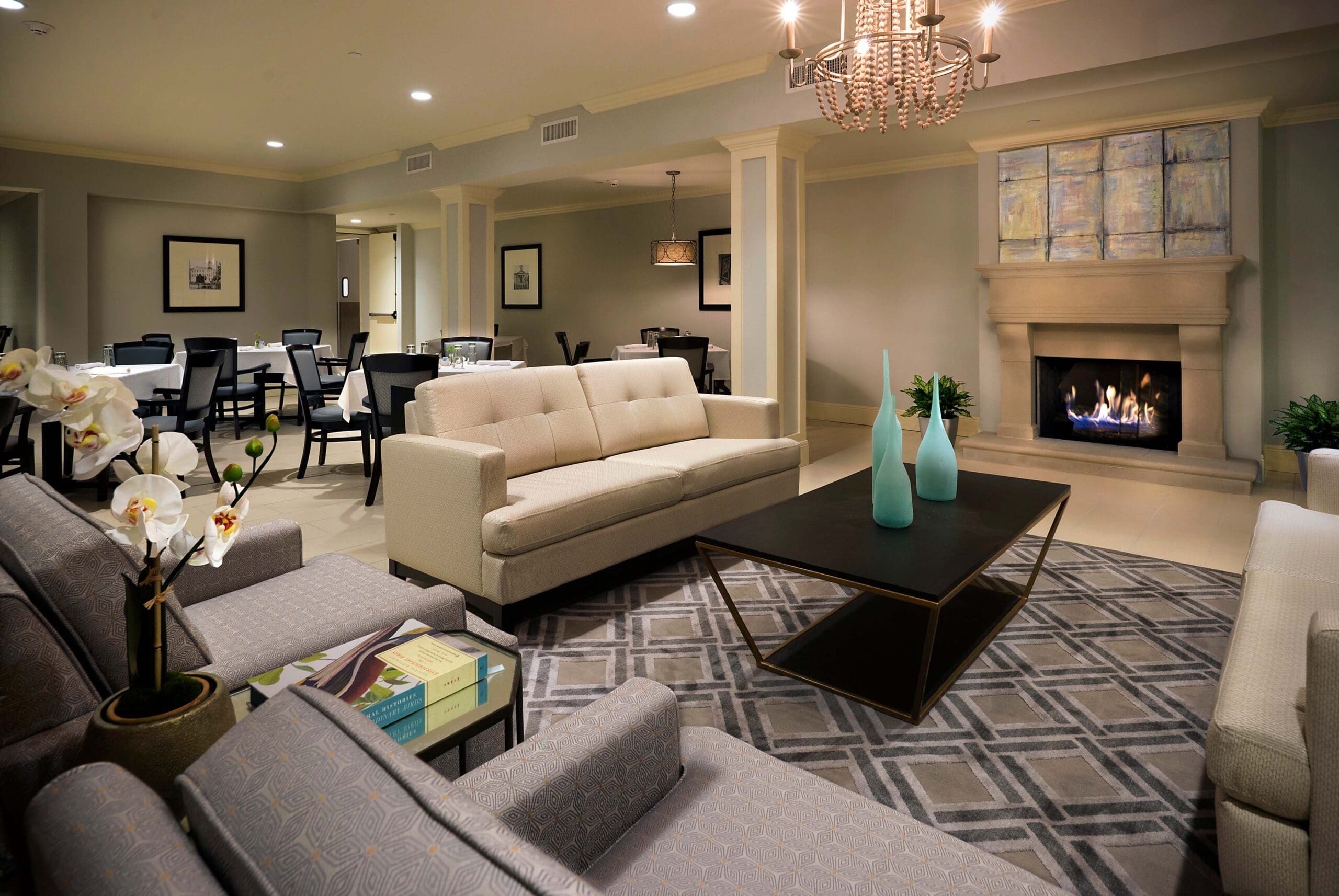 A communal sitting room with a white couch, gray sofa chairs, a coffee table with three turquoise vases, and a digital fire place.