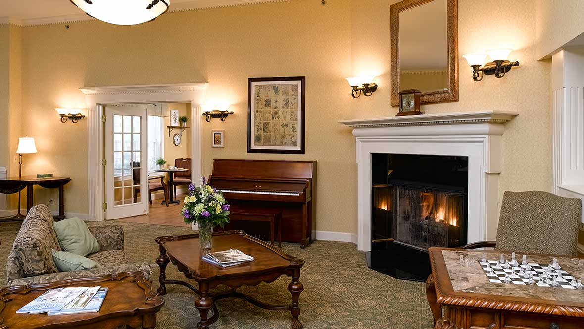 - Grace Management A sitting room with a fireplace, a piano, a couch, two brown coffee tables, a bouquet of flowers inside a vase placed on one of them, and a sofa chair in front of a table with a game of chess on it at Shorehaven, a top choice for senior living near Warren.
