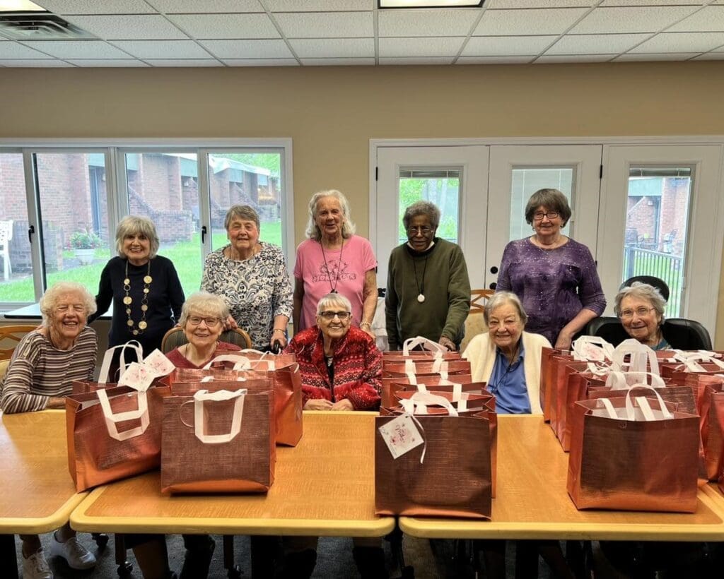 Group of elderly women gathered around a table with red gift bags, smiling at a community event.