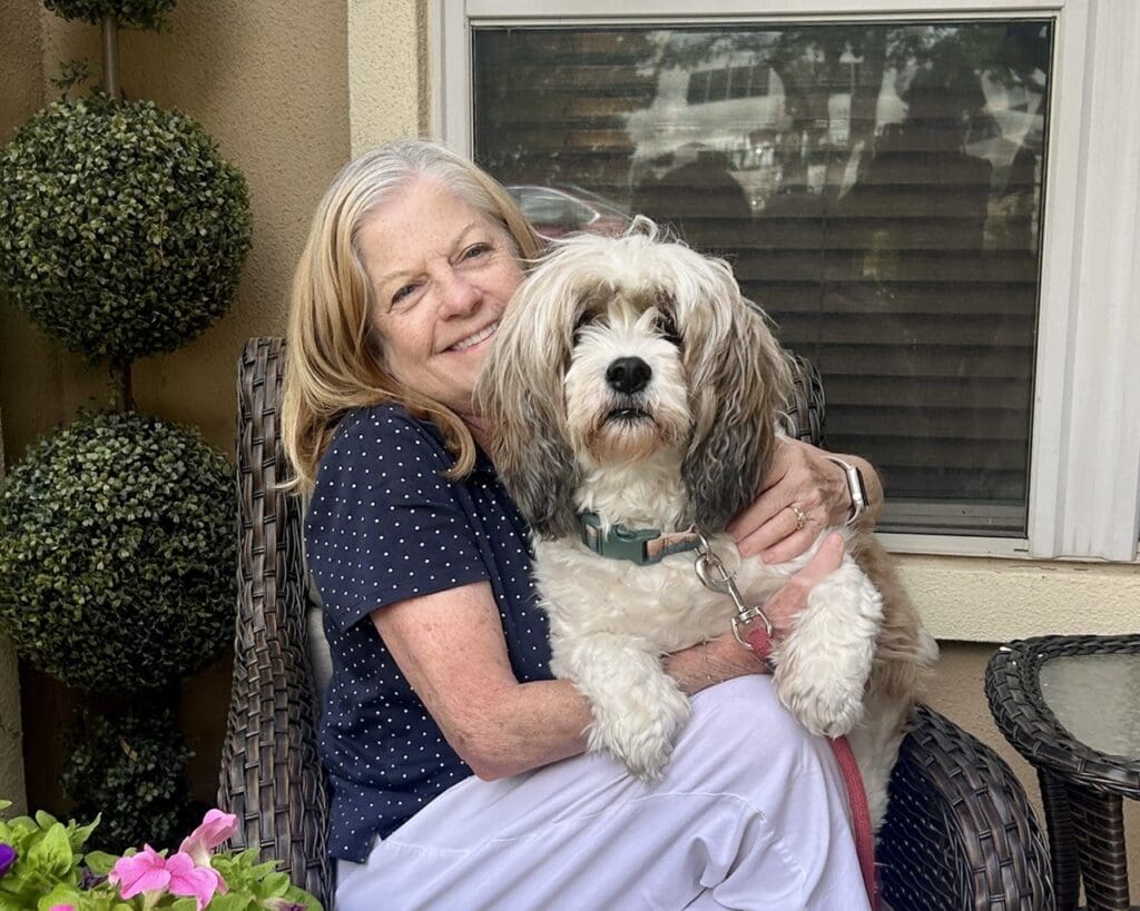 Smiling older woman in a navy polka-dot shirt sits on a wicker chair, hugging a fluffy white-and-gray dog outdoors by a window.