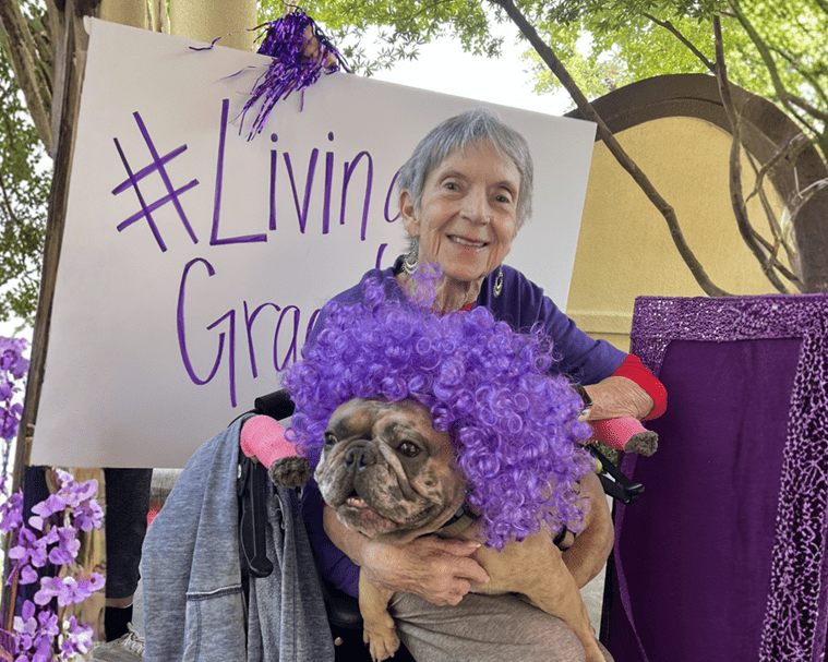 Smiling elderly woman in a wheelchair hugging a bulldog wearing a bright purple curly wig at an outdoor purple-themed event, with a white sign in the background reading '#Living Grace'.