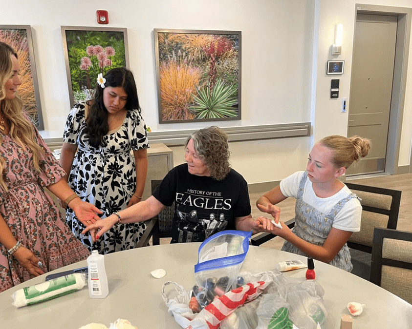 Four people sit and stand around a round table, with nail polish supplies and bags, as someone paints another persons nails and others watch.