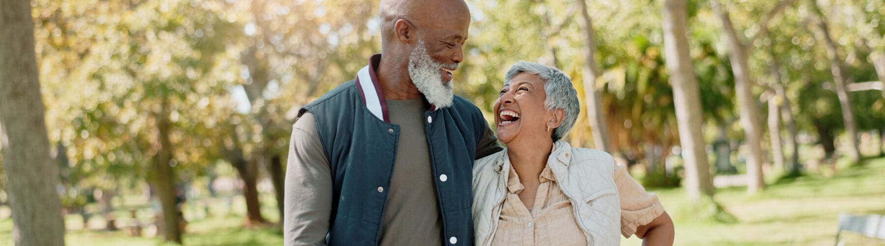 A cheerful couple shares a joyful moment in a sunlit park, surrounded by vibrant greenery and trees.