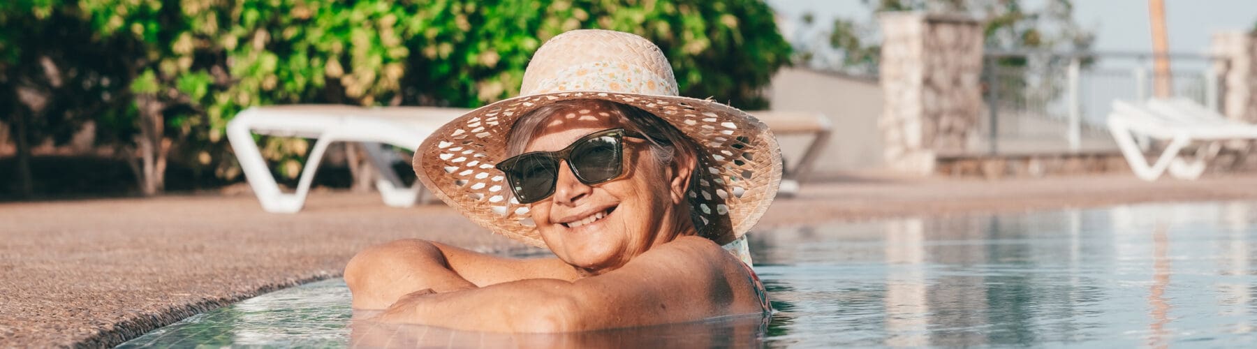 Smiling senior woman in a straw hat enjoys a serene moment in a sparkling pool, surrounded by lush greenery.