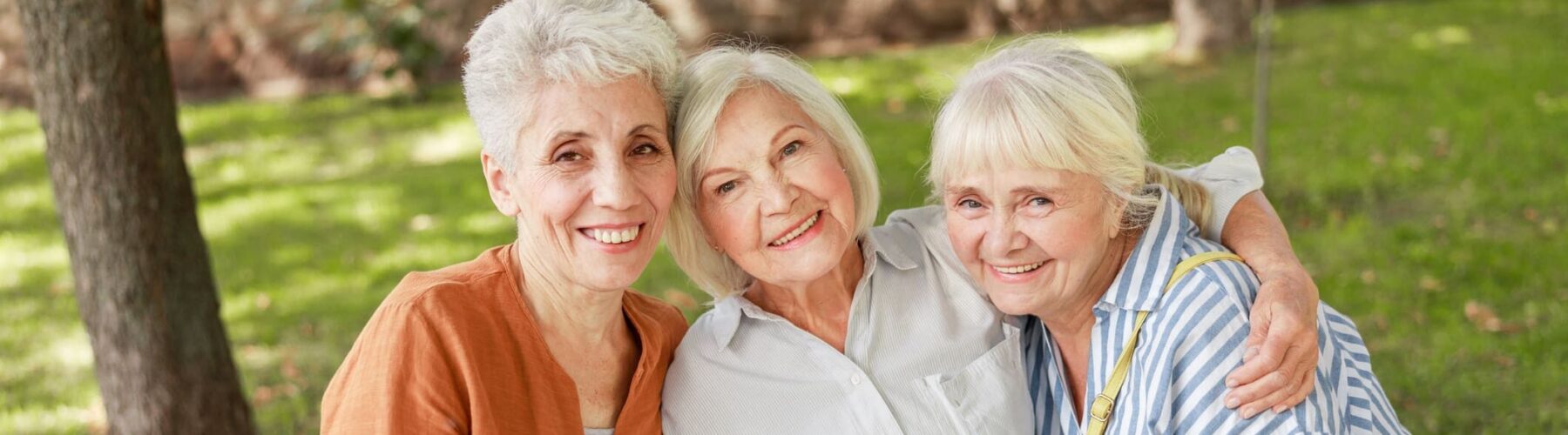 Three cheerful senior women enjoy a sunny day together in a lush green park, sharing smiles and warm companionship.