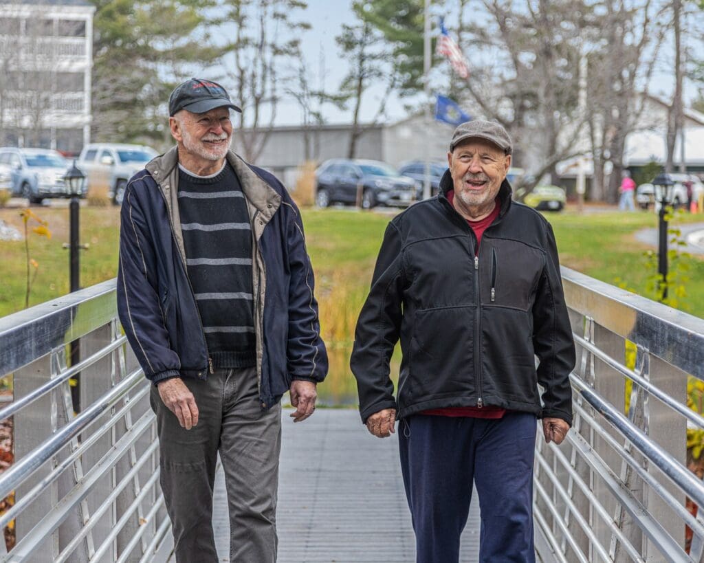 Two elderly men walk side by side on a metal pedestrian bridge, smiling with a park and cars in the background.