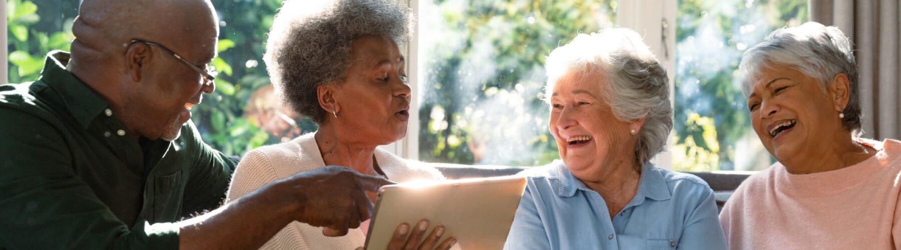 Four seniors on a sofa laugh together while sharing a tablet device in a sunlit living room.