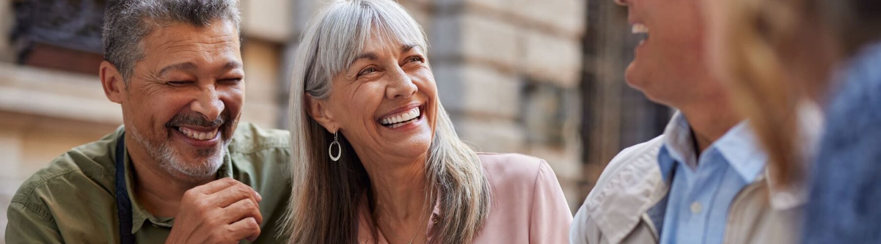 Four adults share a laugh outdoors, center woman with gray hair and sunglasses hanging from her blouse.