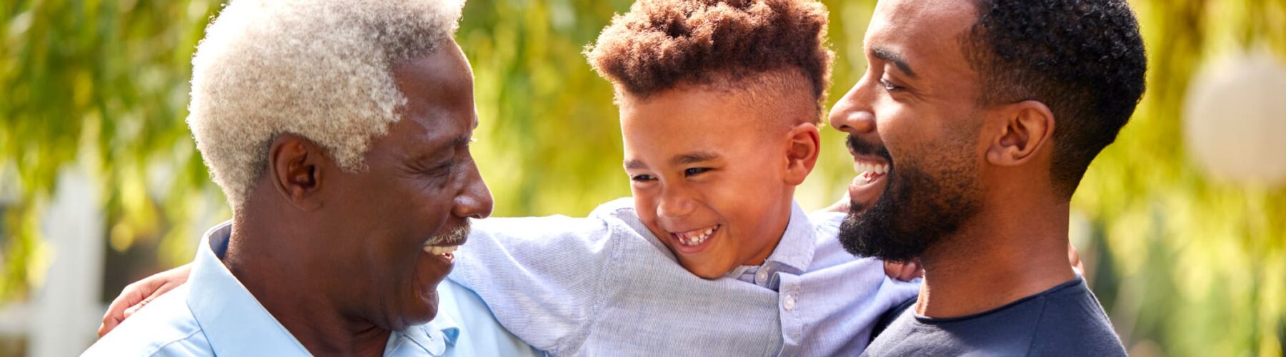 Three generations of men joyfully interact outdoors, sharing smiles in a lush, vibrant setting filled with greenery.