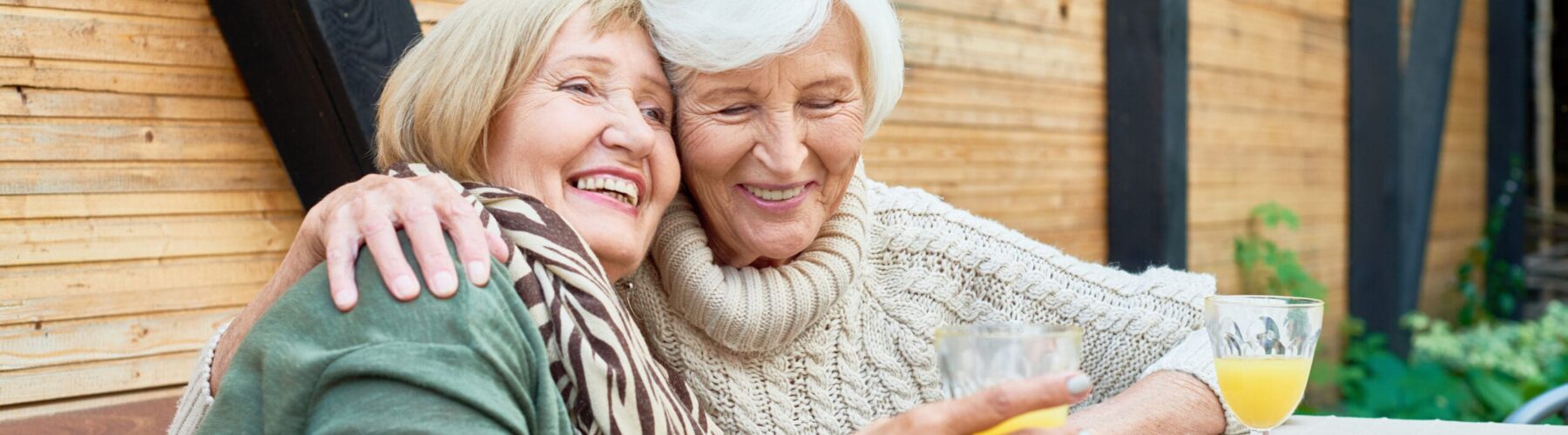 Two senior women share a joyful moment, embracing at a table with drinks and apples in a warm, inviting outdoor setting.