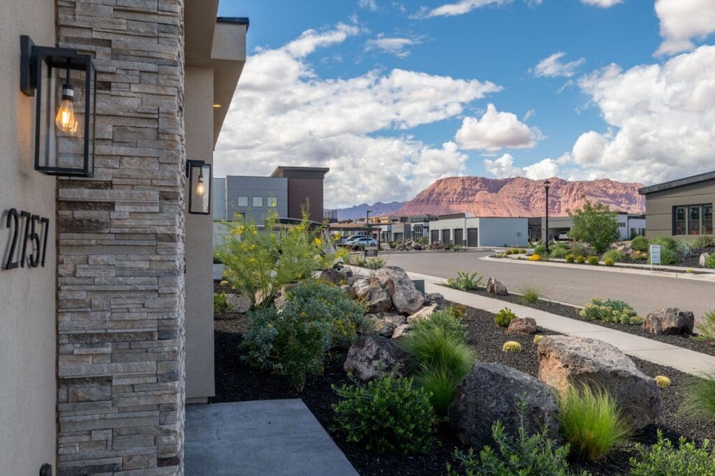 Contemporary senior living community entrance with landscaped gardens and stunning mountain backdrop under a bright sky.