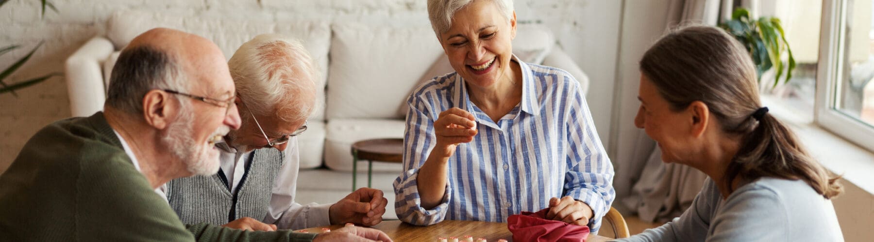 Four seniors joyfully engaging in a lively card game around a wooden table in a bright, inviting living space.