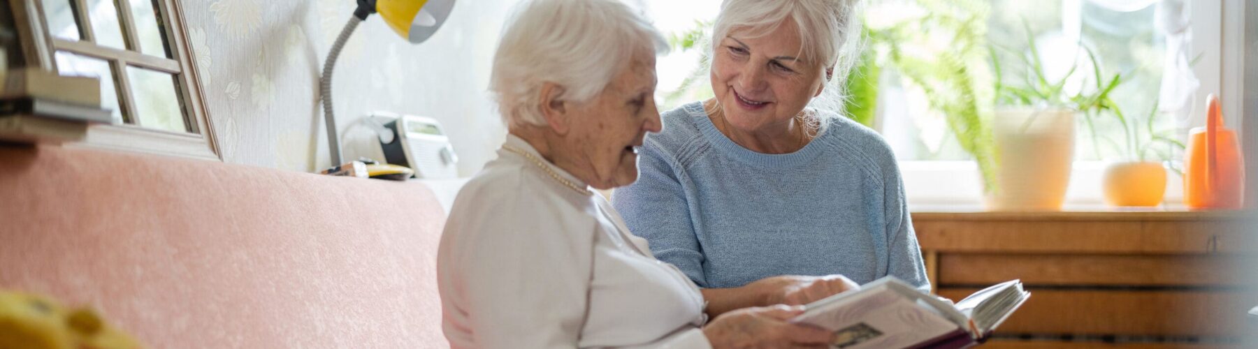 Two senior women share a joyful moment while looking at a photo book together in a bright, inviting living room.